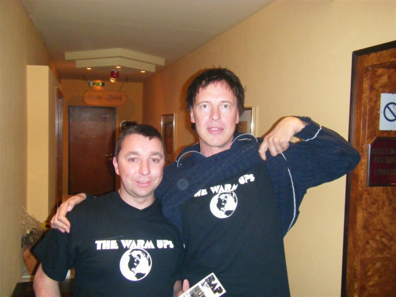 Zwei Männer in schwarzen T-Shirts mit der Aufschrift ‚THE WARM UPS‘ posieren lächelnd in einem Hotelflur.
