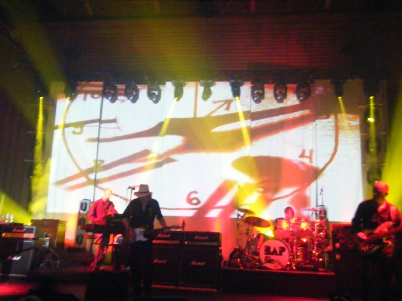 BAP live auf der Bühne während der "Radio Pandora"-Tour 2008/09. Die Band spielt vor einer großen Leinwand mit abstrakten, dynamischen Lichtprojektionen. Im Vordergrund sind Musiker mit Instrumenten wie Schlagzeug, Keyboard und Gitarren zu sehen, umgeben von professioneller Bühnenbeleuchtung.