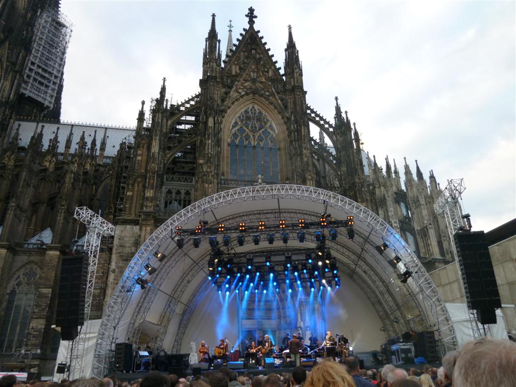 Bühne mit Band vor dem Kölner Dom bei Tageslicht. Die Bühne ist mit Scheinwerfern und einer weißen Überdachung ausgestattet. Im Hintergrund ragt die gotische Fassade des Doms mit ihren Türmen und Fenstern auf. Vor der Bühne steht ein Publikum.