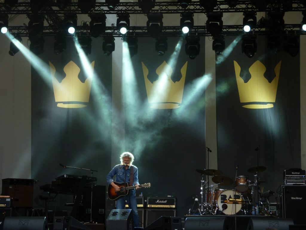Ein Musiker mit grauen Haaren und blauem Hemd spielt eine braune E-Gitarre auf einer dunklen Bühne. Hinter ihm sind zwei große gelbe Kronen-Symbole an der Wand projiziert. Spotlights erhellen die Szene, während im Hintergrund ein Schlagzeug und Verstärker zu sehen sind.
