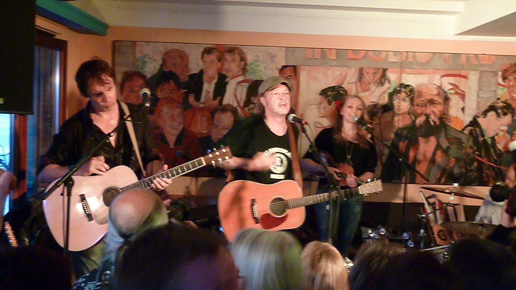 Drei Musiker spielen auf einer kleinen Bühne vor Publikum. Beide Musiker halten helle Akustikgitarren, die Musikerin eine kleine akustische Gitarre. Der mittlere Musiker singt i ein Mikrofon. Im Hintergrund sind weitere Personen auf einem Wandgemälde zu sehen.