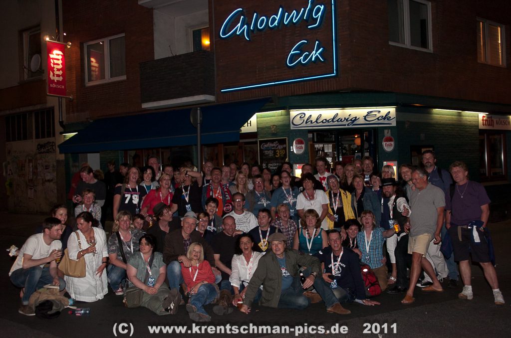 Eine große Gruppe von etwa 50 Personen posiert vor dem Eingangsbereich des Lokals "Chlodwig Eck" in Köln. Die Fassade ist mit einer blauen Neon-Schrift "Chlodwig Eck" beleuchtet. Viele tragen Lanyards und BAP-Shirts.