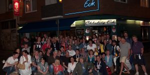 Eine große Gruppe von etwa 50 Personen posiert vor dem Eingangsbereich des Lokals "Chlodwig Eck" in Köln. Die Fassade ist mit einer blauen Neon-Schrift "Chlodwig Eck" beleuchtet. Viele tragen Lanyards und BAP-Shirts.