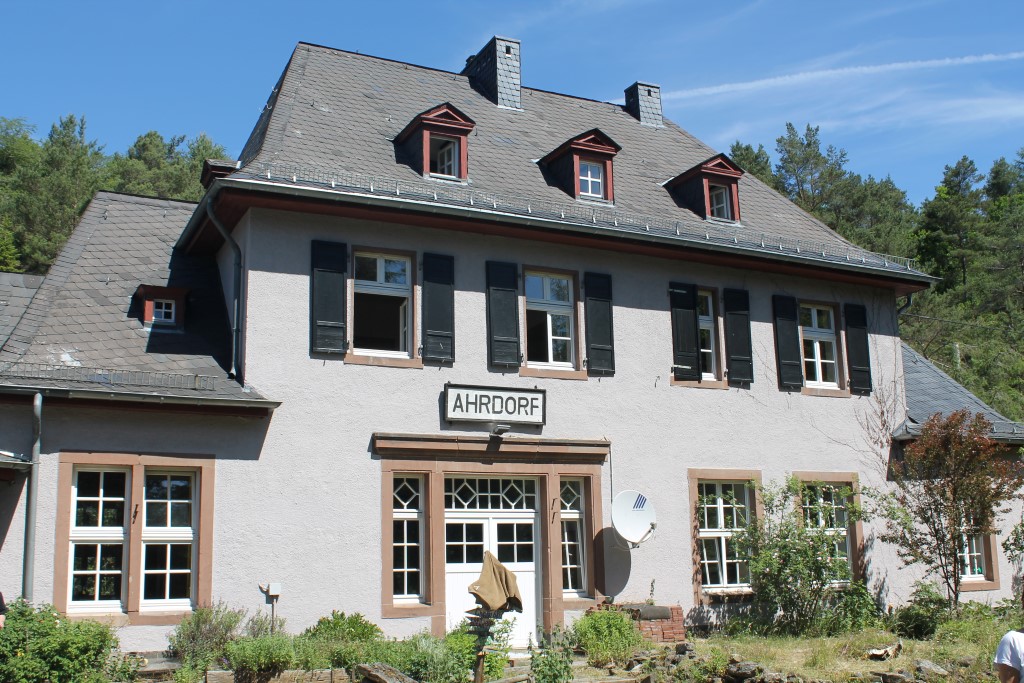Ein zweistöckiges Haus mit hellem Putz, dunklem Schieferdach und mehreren Dachgauben. Über dem Haupteingang hängt ein Schild mit der Aufschrift „AHRDORF“. Das Haus ist von Grünpflanzen und Bäumen umgeben, im Hintergrund sind weitere Bäume zu sehen.