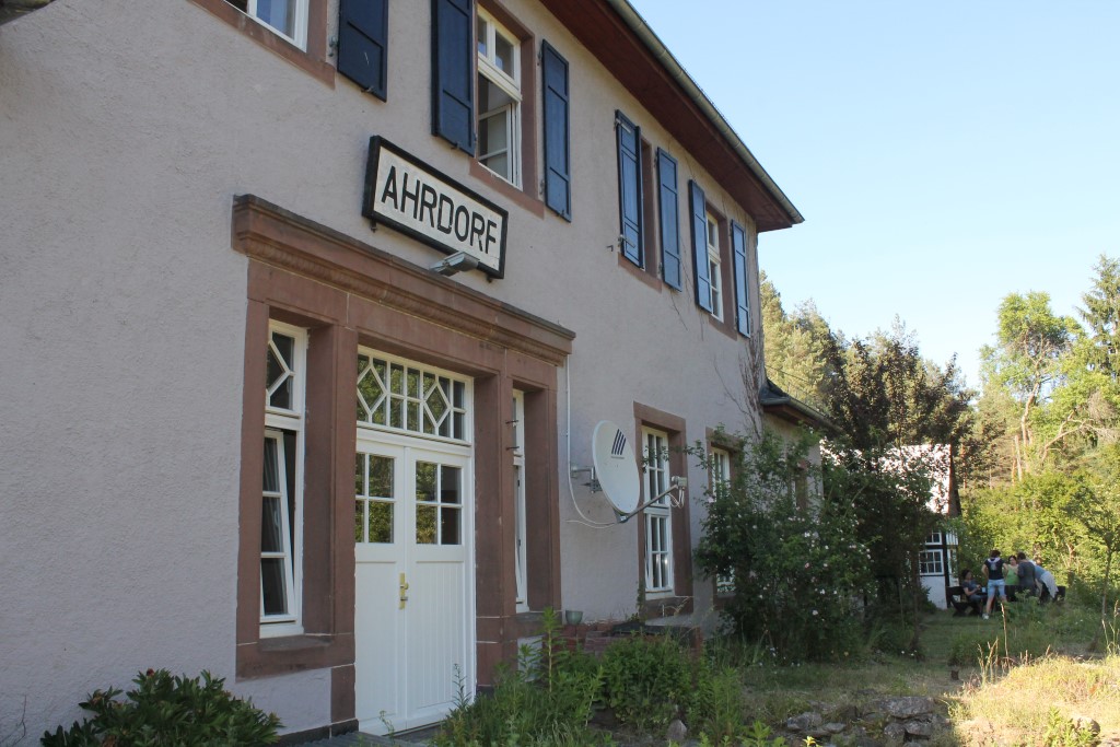 Ein zweistöckiges Haus mit hellem Putz, braunen Fensterrahmen und einem Schild mit der Aufschrift „AHRDORF“ über dem Eingang. Neben dem Haus wächst ein Strauch, und im Hintergrund sind Bäume sowie eine Person im Garten zu sehen.