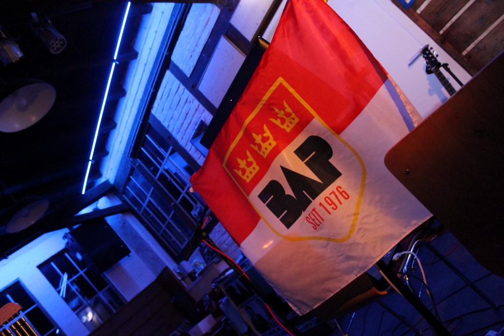 Eine rot-weiße Flagge mit dem Logo „BAP“ und dem Schriftzug „seit 1976“ in einem Raum mit blauer Beleuchtung. Im Hintergrund ist eine Fachwerkwand zu erkennen.