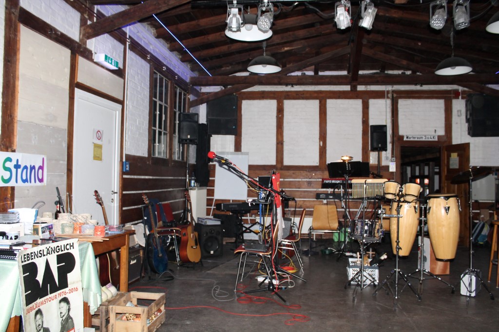 Ein Proberaum mit Holzwänden, einer Holzdecke und mehreren Musikinstrumenten. Im Vordergrund stehen Percussions, Congas und ein Mikrofonständer. An der Wand hängt ein Plakat mit der Aufschrift „Lebenslänglich BAP Jubiläumstour 1976-2016“. Links im Raum sind Gitarren und weitere Ausrüstung zu sehen.