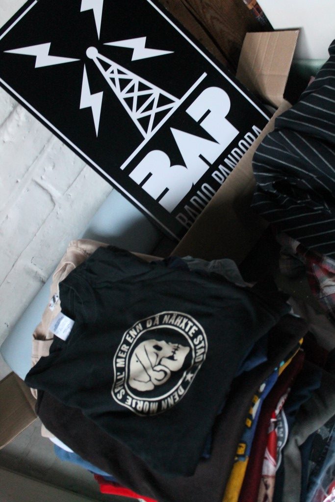 Ein schwarzes T-Shirt mit einem Aufdruck, der einen Totenschädel und die Worte „Denn Morje spill' mer enn dä Nähxte Stadt“ zeigt. Daneben liegt ein schwarzes Schild mit der Aufschrift „BAP Radio Pandora“ und einem Blitzsymbol.