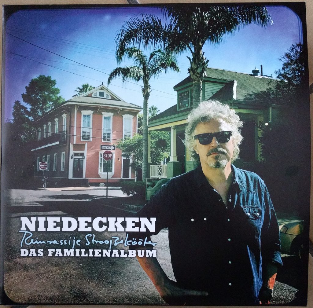 Ein Albumcover mit dem Titel "NIEDECKEN – Das Familienalbum" und dem Untertitel "Reinrassige Strooßeköter". Im Hintergrund ist ein rotes Haus mit weißer Veranda und Palmen zu sehen. Im Vordergrund steht ein Mann mit grauen Haaren, Bart und Sonnenbrille, der ein blaues Hemd trägt.