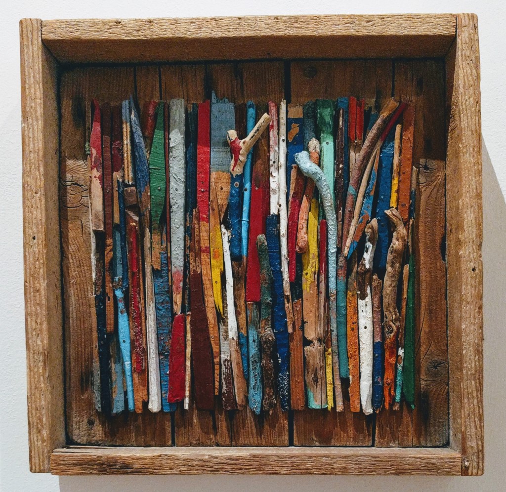 Ein Holzrahmen mit vertikalen, farbig bemalten Holzstäben in verschiedenen Farben wie Rot, Blau, Grün, Gelb und Weiß. Einige Stäbe sind natürlich belassen oder haben eine raue Oberfläche.