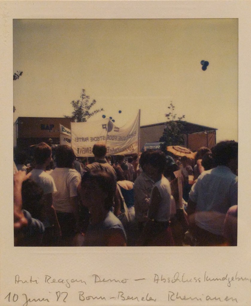 Ein Polaroidfoto zeigt eine Gruppe von Menschen bei einer Demonstration mit einem Transparent. Im Hintergrund sind Bäume, eine Bühne und ein LKW mit der Aufschrift "BAP" zu sehen. Unten steht handschriftlich: "Anti-Reagan-Demo – Abschlusskundgebung - 10. Juni 82 Bonn-Beueler Rheinauen".