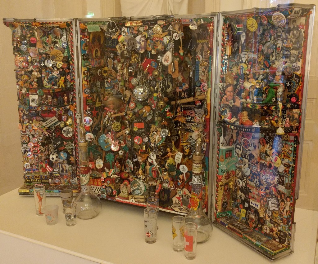Ein dreiteiliger, offener Schrank, der vollständig mit Aufklebern, Buttons, Fotos, kleinen Figuren und anderen Sammelstücken bedeckt ist. Vor dem Schrank stehen mehrere leere Gläser und zwei Flaschen.