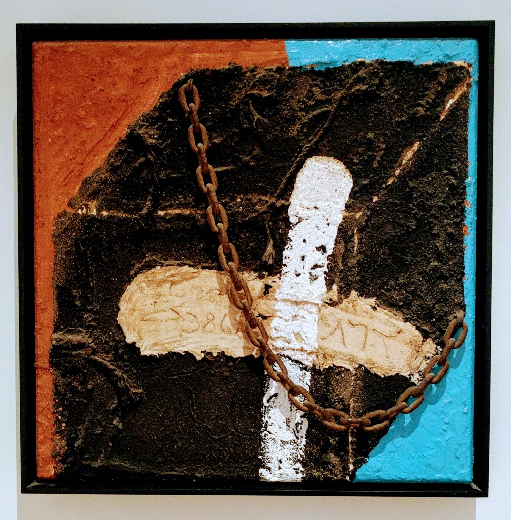 Ein abstraktes Gemälde mit einer rostigen Kette, die diagonal über die Leinwand verläuft. Im Hintergrund sind dunkle, erdige Farben, ein weißer Streifen und ein beigefarbener Bereich zu sehen. Links oben ist ein oranger Farbakzent, rechts unten ein blauer Bereich.