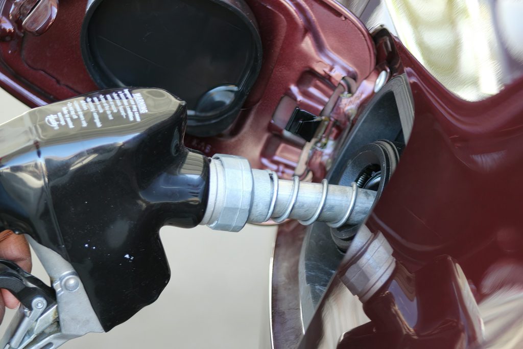 Eine Hand hält einen schwarzen Zapfgriff einer Tankstelle und betankt ein rotes Auto. Im Hintergrund sind Teile des Tankstutzen und der Karosserie sichtbar.