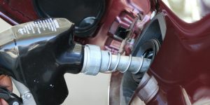 Eine Hand hält einen schwarzen Zapfgriff einer Tankstelle und betankt ein rotes Auto. Im Hintergrund sind Teile des Tankstutzen und der Karosserie sichtbar.