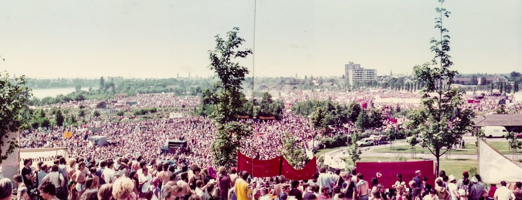 Panoramaaufnahme einer großen Menschenmenge auf einer weiten Grünfläche bei einem Open-Air-Event. Im Vordergrund stehen zahlreiche Menschen in sommerlicher Kleidung, einige mit bunten Accessoires. Im Hintergrund sind Bäume, Gebäude und eine leicht hügelige Landschaft zu erkennen. Die gedämpften Farbtöne deuten auf eine ältere Aufnahme hin.