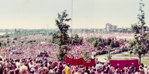 Panoramaaufnahme einer großen Menschenmenge auf einer weiten Grünfläche bei einem Open-Air-Event. Im Vordergrund stehen zahlreiche Menschen in sommerlicher Kleidung, einige mit bunten Accessoires. Im Hintergrund sind Bäume, Gebäude und eine leicht hügelige Landschaft zu erkennen. Die gedämpften Farbtöne deuten auf eine ältere Aufnahme hin.
