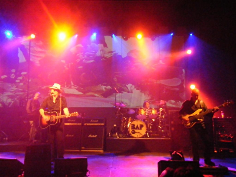 BAP live auf der Bühne, beleuchtet in Rot, Blau und Gelb. Im Vordergrund ein Mann mit Hut an der Gitarre, daneben der Bassist, im Hintergrund Schlagzeuger und Keyboarder.