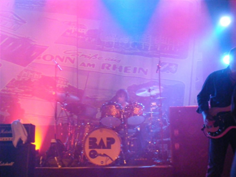 Bühne mit Schlagzeug, auf der Bassdrum das BAP-Logo, dahinter ein Banner mit der Aufschrift "BONN AM RHEIN". Blau-rotes Licht, rechts ein Bassist teilweise zu sehen.